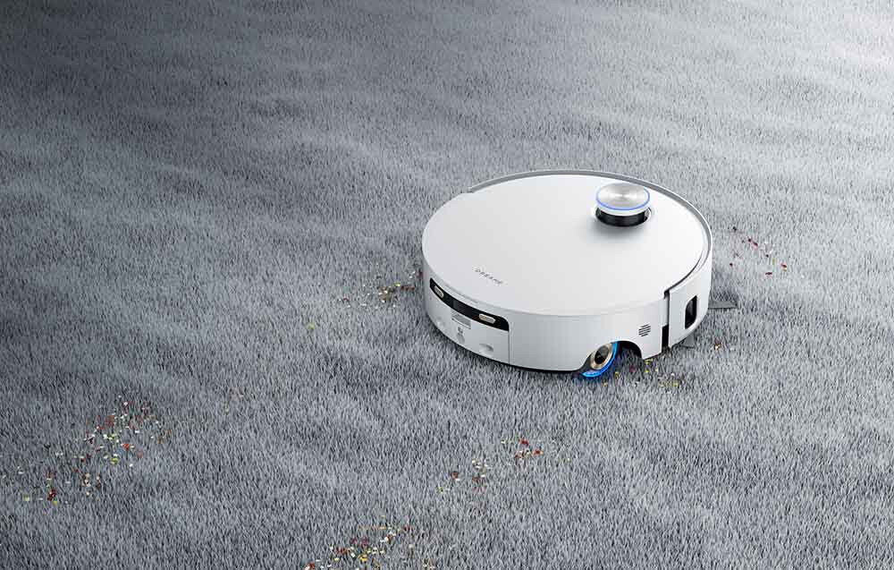 Biały robot sprzątający porusza się po szarej powierzchni dywanu, zbierając drobne kolorowe elementy. Na górze urządzenia widoczny jest okrągły element, a na boku znajduje się logo producenta.