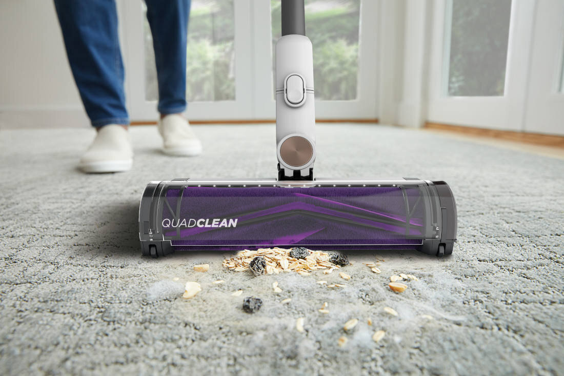 Odkurzacz z napisem \'QUADCLEAN\' odkurza dywan, zbierając rozsypane okruchy i kurz. W tle widoczne są nogi osoby stojącej na dywanie oraz duże okna.