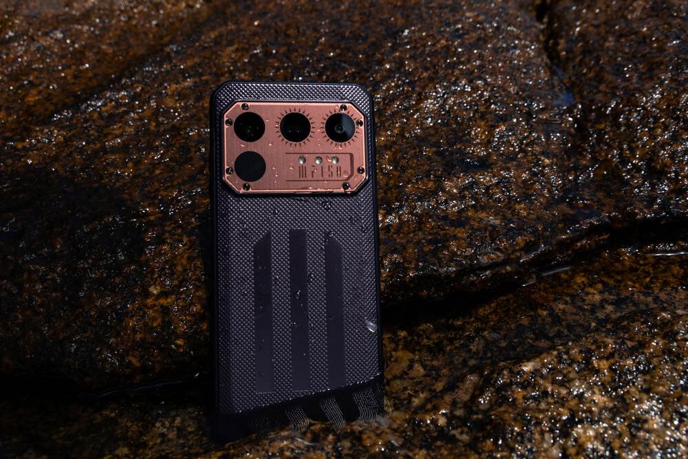 Smartfon z widocznym aparatem i metaliczną płytką z napisem \'IIIF150\' stoi na mokrej, kamienistej powierzchni.