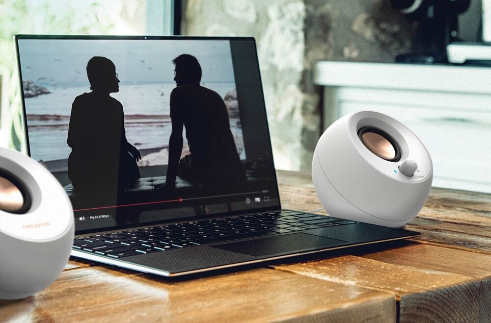 Na drewnianym stole stoi laptop z wyświetlonym obrazem dwóch sylwetek siedzących nad wodą, obok znajdują się dwa białe głośniki o kulistym kształcie z widocznym logo \'CREATIVE\'.