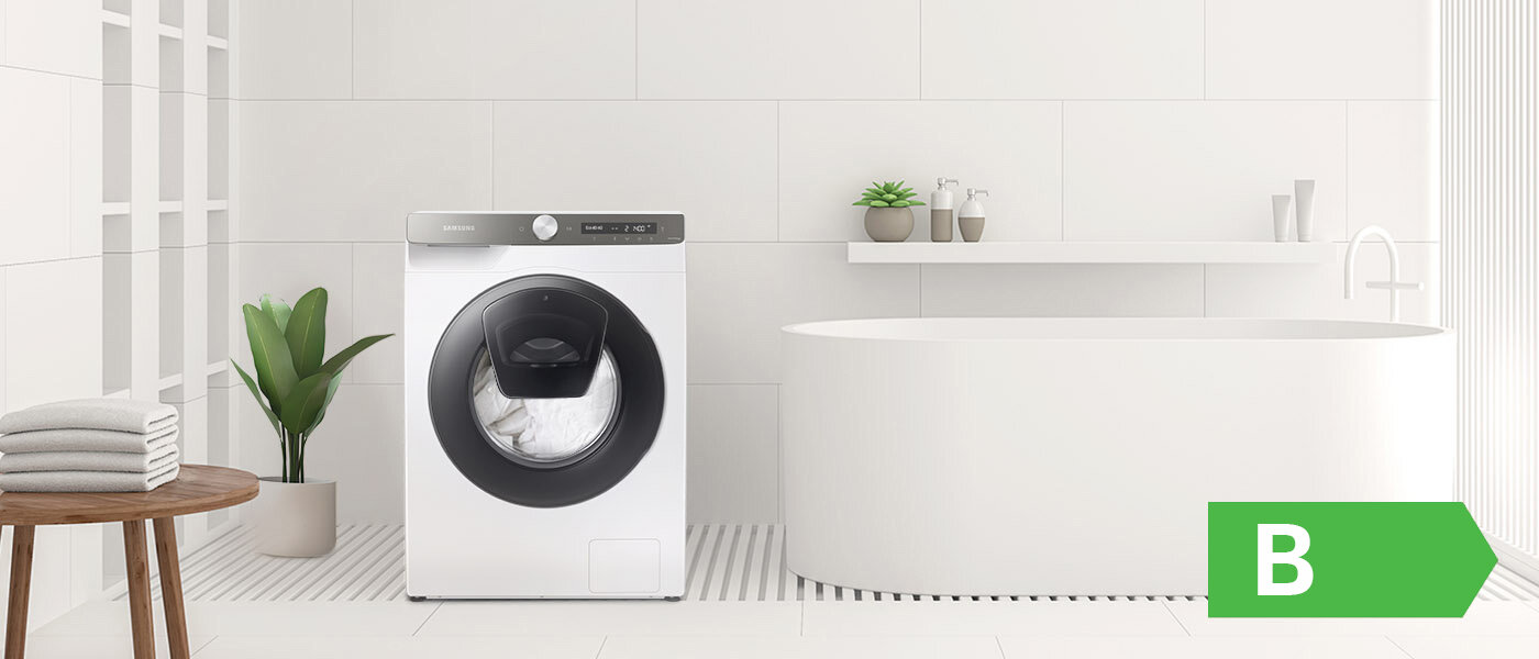 phplL5P75 phpI0Z7sB php6M2kOp WW80T554DAT klasa energetyczna 1400 (N13) W jasnej łazience znajduje się pralka z dużym okrągłym drzwiczkami, obok której stoi roślina w doniczce. Na prawo od pralki jest wolnostojąca wanna, a w dolnym rogu widoczny jest zielony symbol z literą \'B\'.