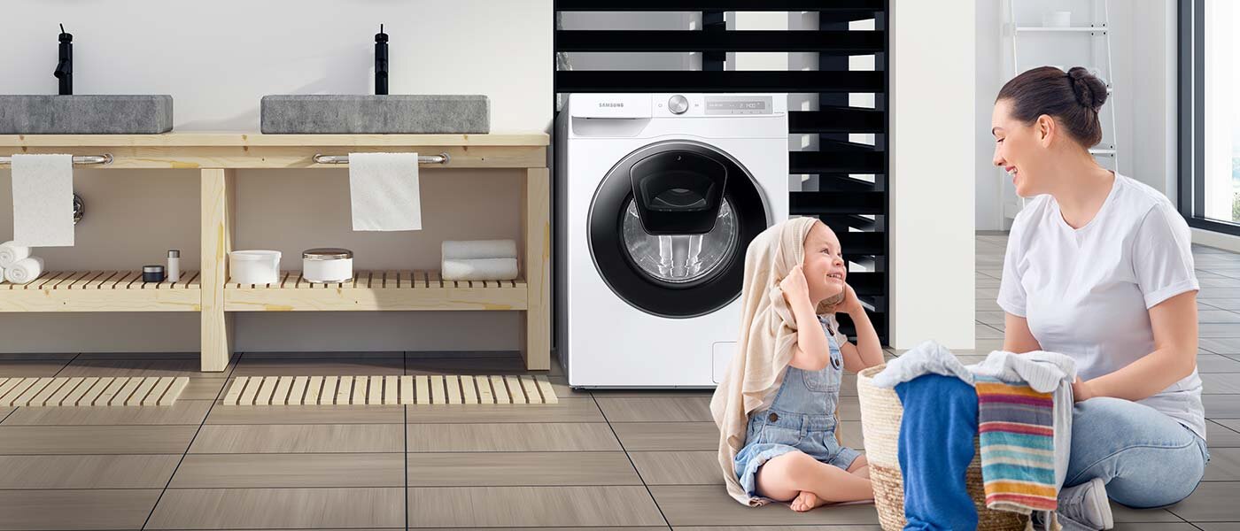 Pralka Samsung w tle, z przodu kobieta z dzieckiem W pomieszczeniu znajduje się pralka z widocznym logo \'SAMSUNG\', obok której siedzą dwie osoby z koszem pełnym prania. W tle widać drewniane półki z umywalkami i ręcznikami.