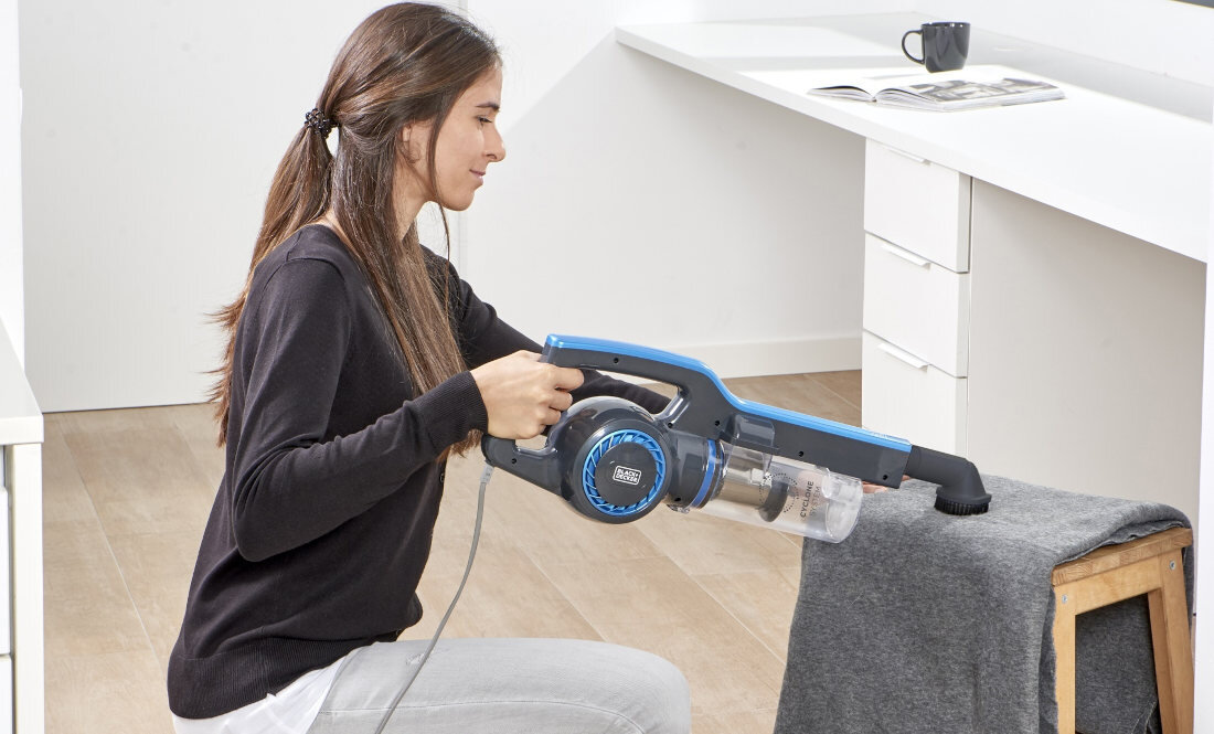 Osoba używa odkurzacza ręcznego BLACK&DECKER do czyszczenia materiału na stołku. W tle znajduje się biurko z otwartą książką i kubkiem.
