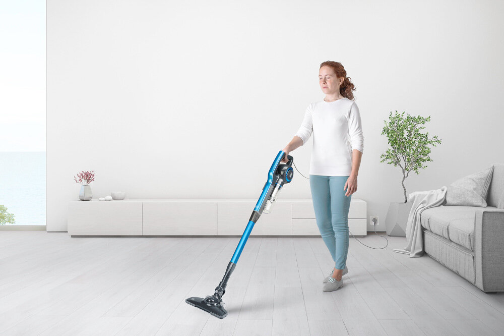 Osoba używa niebieskiego odkurzacza w jasnym, minimalistycznym wnętrzu z sofą i rośliną w tle.