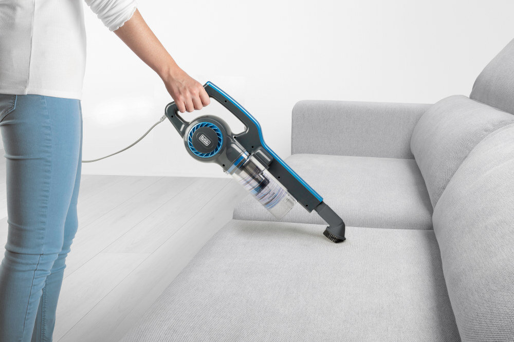 Osoba używa odkurzacza ręcznego z niebieskimi akcentami do czyszczenia jasnej kanapy. Na odkurzaczu widoczne jest logo BLACK&DECKER.