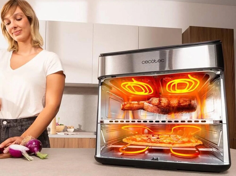 Na blacie kuchennym stoi frytkownica beztłuszczowa Cecotec z widocznym wnętrzem, w którym pieczone są mięso i pizza. Obok urządzenia znajduje się osoba oraz kilka warzyw, w tym cebula i czosnek.