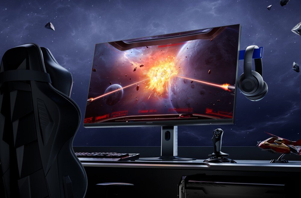 Monitor XIAOMI G Pro 27Qi Stanowisko do gier komputerowych z monitorem wyświetlającym wybuch w kosmosie. Nad monitorem visi czarny hełm. Z lewej strony monitory visi czarne słuchawki. Z prawej strony visi czarny fotel, HDR, jasność, kontrast