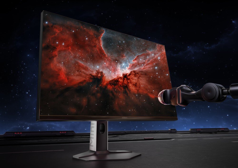 Monitor XIAOMI G Pro 27Qi Monitor z ekranem skierowanym w lewą stronę, wspierany przez czarne mechaniczne ramię. Ekran pokazuje czerwoną i niebieską mgławicę z wieloma gwiazdami. Monitor stoi na czarnym, gładkim podłożu z podłużnymi listwami świetlnymi, kolory, odwzorowanie barw, dokładność