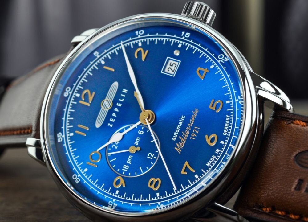 Zegarek z niebieską tarczą, złotymi cyframi i wskazówkami, z logo \'ZEPPELIN\' oraz napisami \'automatic\', \'Mediterranee\', \'1921\', \'MADE IN GERMANY\'. Na tarczy widoczny jest datownik z liczbą \'25\'.
