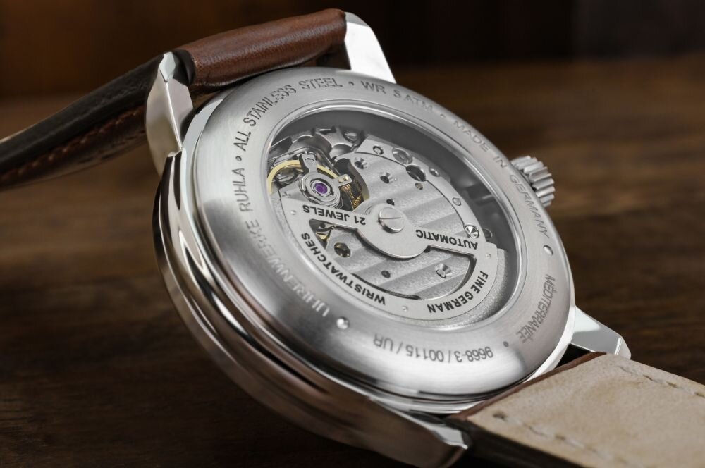 Zbliżenie na tylną część zegarka z widocznym mechanizmem automatycznym i napisem \'ALL STAINLESS STEEL - WR 5 ATM - AUTOMATIC - 21 JEWELS\'. Pasek zegarka jest wykonany z jasnej skóry.