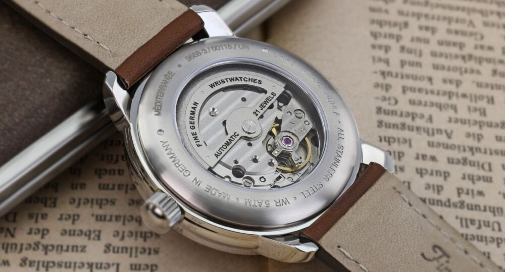Zbliżenie na tylną część zegarka z widocznym mechanizmem i napisem: \'FINE GERMAN WRISTWATCHES, AUTOMATIC, 21 JEWELS, MADE IN GERMANY, ALL STAINLESS STEEL, WATER RESISTANT\'.