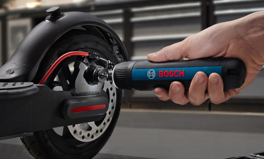 Osoba używa wkrętaka akumulatorowego marki BOSCH do pracy przy kole hulajnogi elektrycznej. Wkrętak jest czarny z niebieskim panelem i czerwonym logo BOSCH.