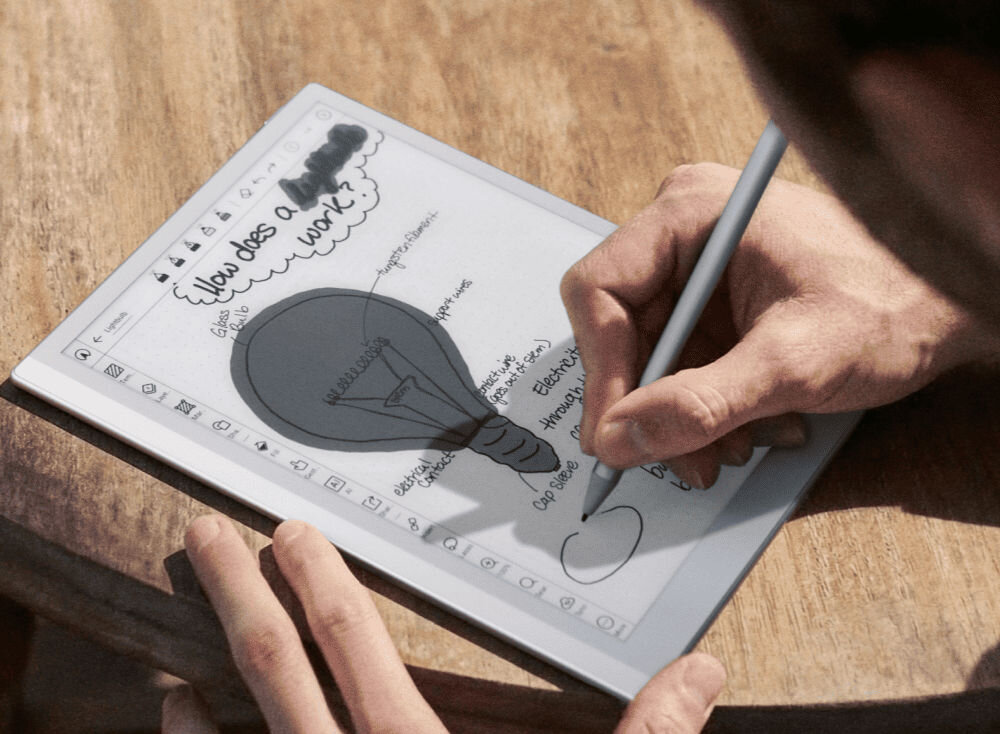 Osoba pisze rysikiem na ekranie czytnika e-booków, na którym widoczny jest szkic żarówki i notatki. Na ekranie znajduje się tekst: \'How does a work?\'.