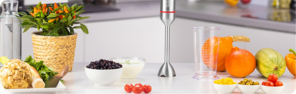 Na blacie kuchennym znajduje się blender ręczny w pobliżu różnych owoców i warzyw, w tym pomarańczy, pomidorów i jagód. Obok blendera stoi przezroczysty pojemnik, a w tle widoczna jest doniczka z rośliną.