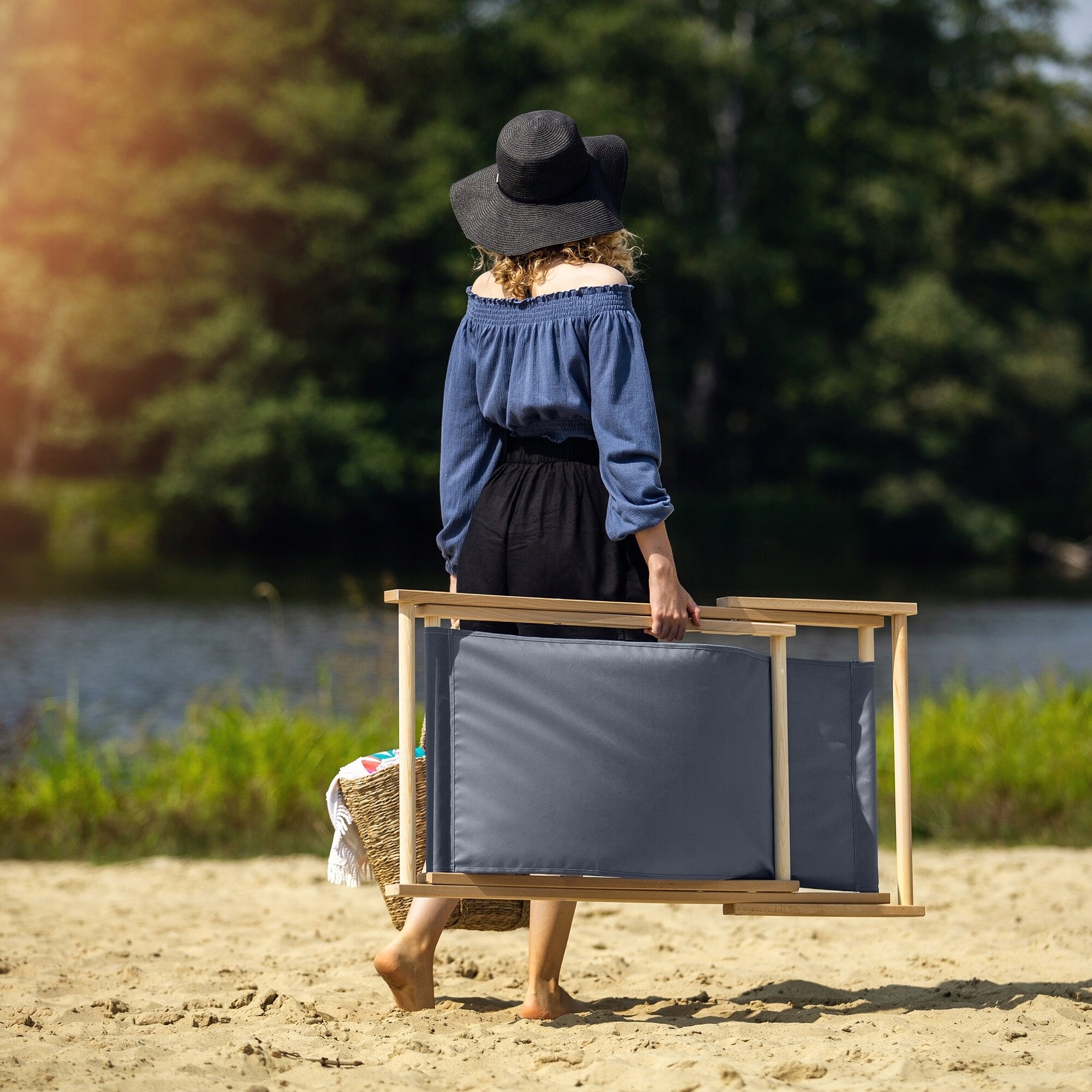 Osoba na plaży niesie składany leżak drewniany z szarym materiałem. W tle widać wodę i drzewa.