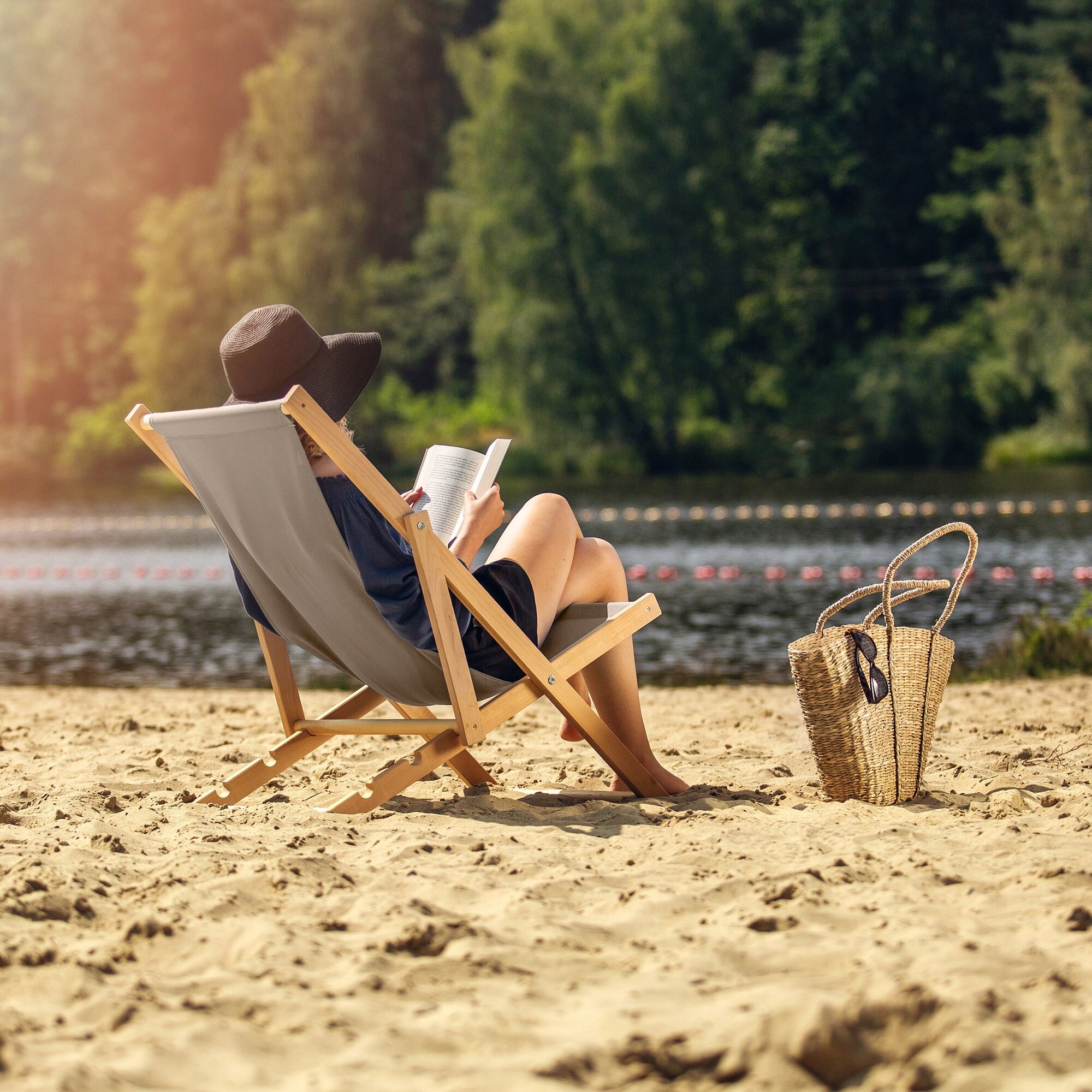 Osoba siedzi na drewnianym, składanym leżaku na piaszczystej plaży, trzymając książkę. Obok leżaka znajduje się pleciona torba z okularami przeciwsłonecznymi.