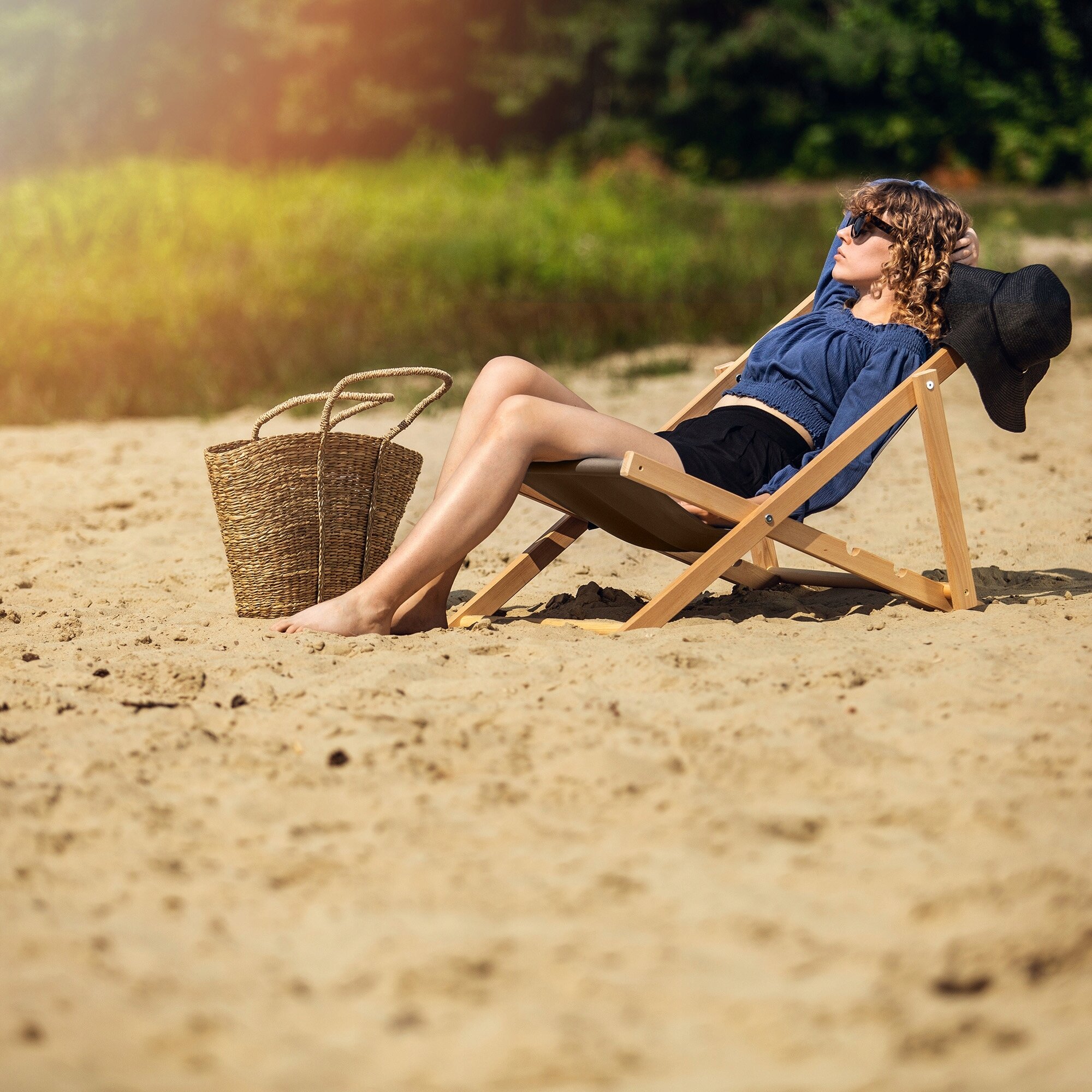 Osoba leży na drewnianym, składanym leżaku na piaszczystej plaży, obok znajduje się pleciona torba. W tle widać zieleń i drzewa.