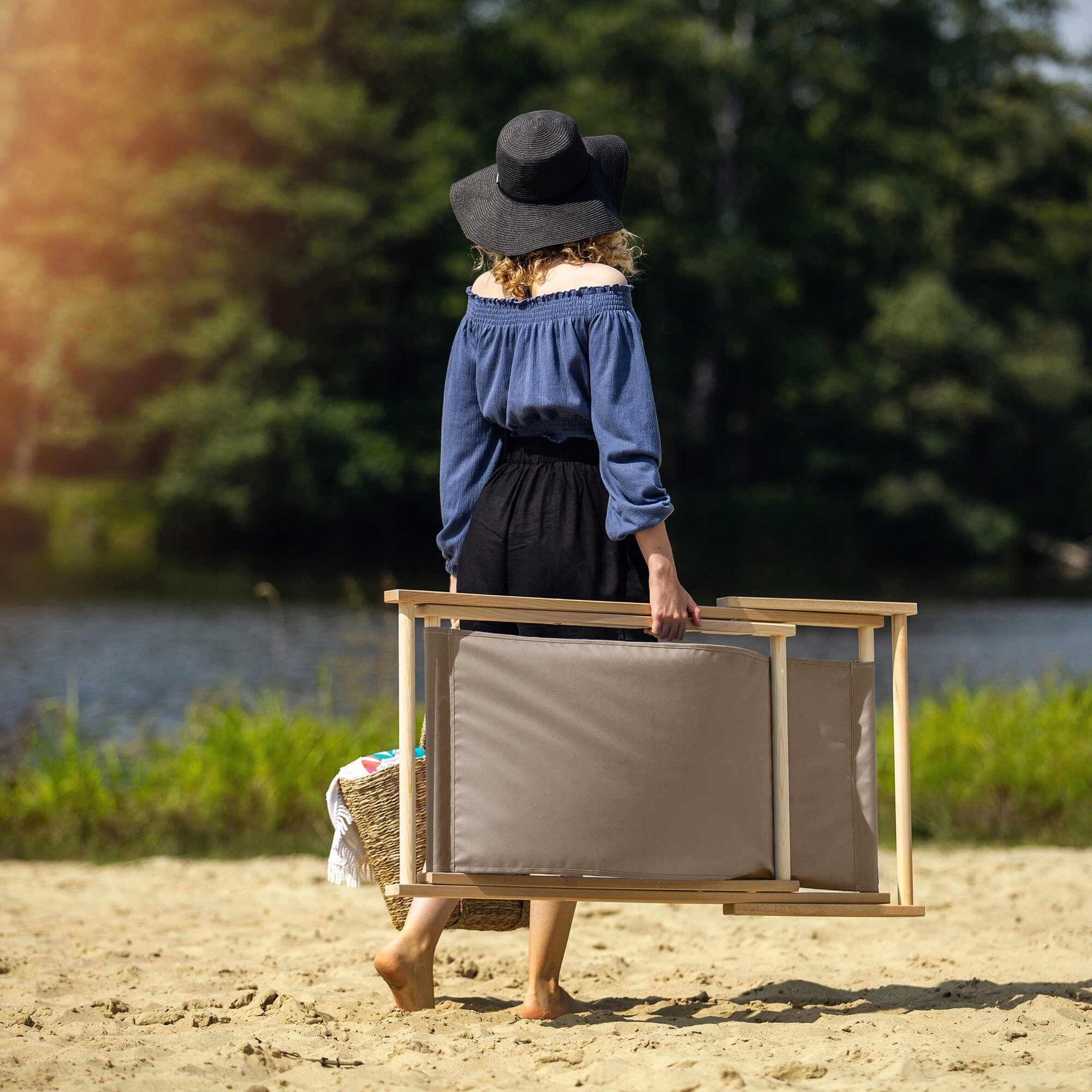 Osoba na plaży niesie składany leżak drewniany. W tle widać wodę i drzewa.