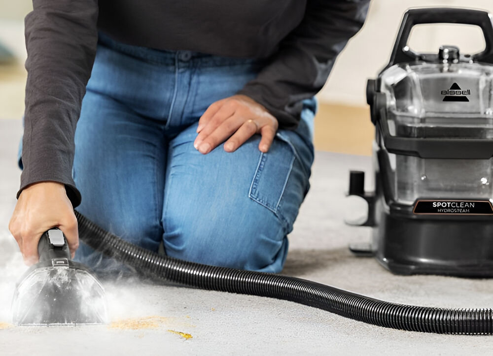 Osoba używa odkurzacza piorącego na dywanie, trzymając w ręku końcówkę ssącą. Na urządzeniu widoczne jest logo \'BISSELL\' oraz napis \'SPOTCLEAN HYDROSTEAM\'.