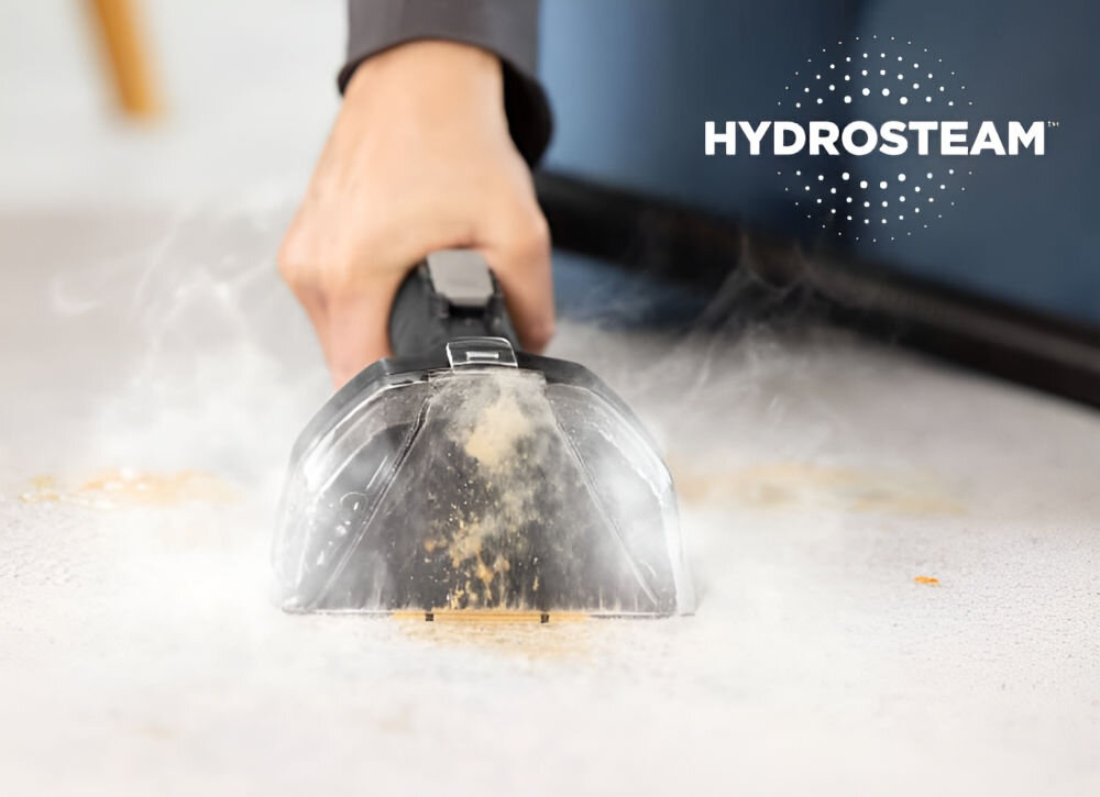 Osoba używa odkurzacza piorącego do czyszczenia dywanu, z którego unosi się para. Widoczny jest napis \'HYDROSTEAM\'.