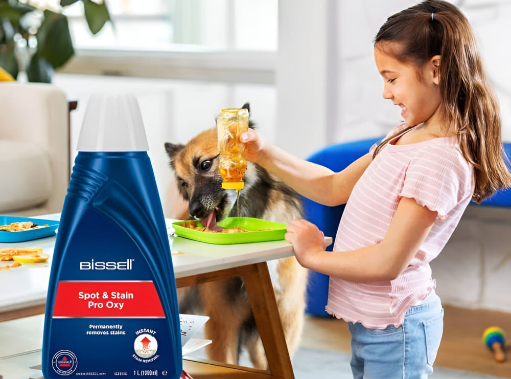 Na pierwszym planie widoczna jest butelka z napisem \'Bissell Spot & Stain Pro Oxy\'. W tle dziecko nalewa płyn z butelki na talerz, a obok stoi pies.