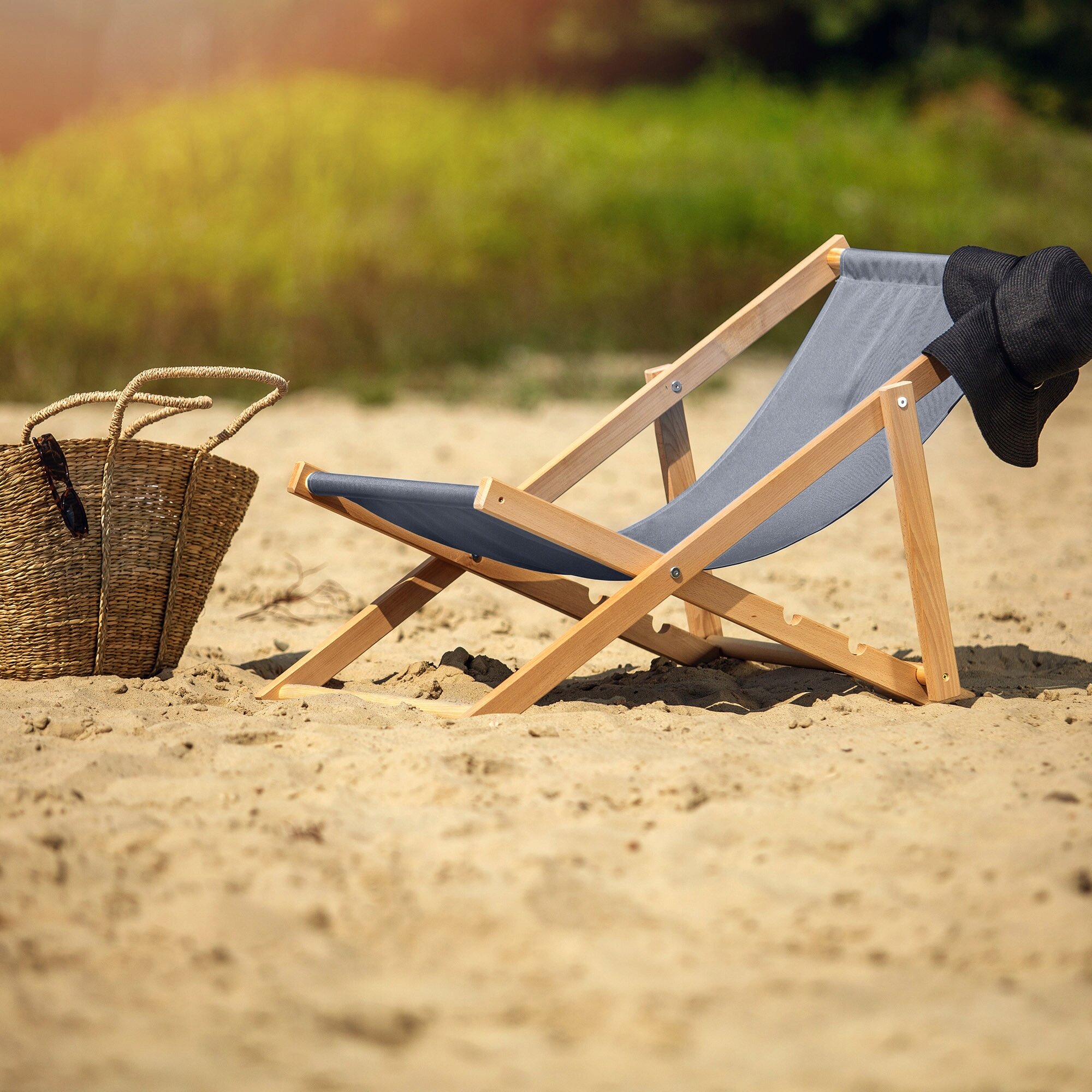 Na piaszczystej plaży znajduje się drewniany leżak z szarym materiałem, obok którego leży czarny kapelusz. Obok leżaka stoi pleciona torba plażowa.