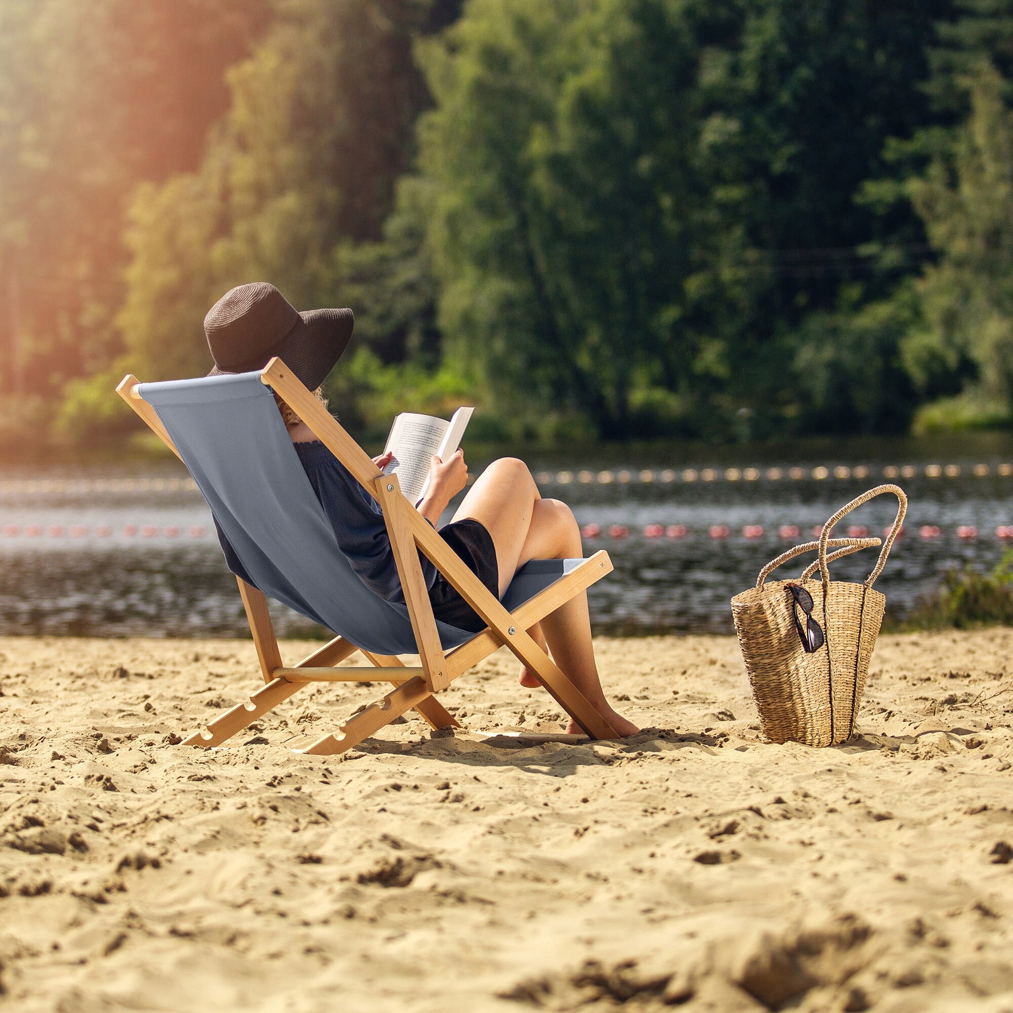 Osoba siedzi na drewnianym leżaku na piaszczystej plaży, trzymając książkę. Obok leżaka znajduje się pleciona torba z okularami przeciwsłonecznymi.