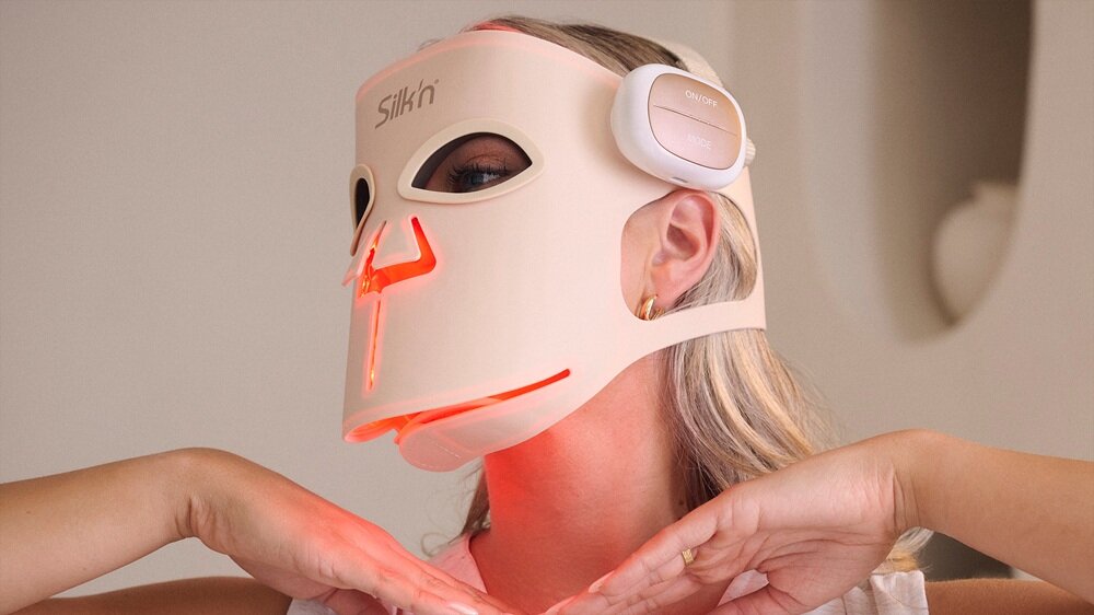 Osoba nosi białą maskę LED na twarzy z widocznym czerwonym światłem i napisem \'Silk\'n\' na górze. Maska ma otwory na oczy i usta oraz dodatkowy element na boku głowy.