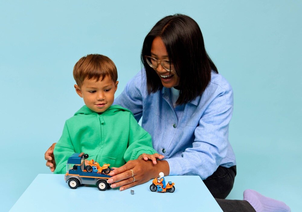 Dwoje ludzi bawi się zestawem LEGO, który zawiera lawetę i motocykle. Tło jest jasnoniebieskie, a stół, na którym znajdują się klocki, jest również niebieski.