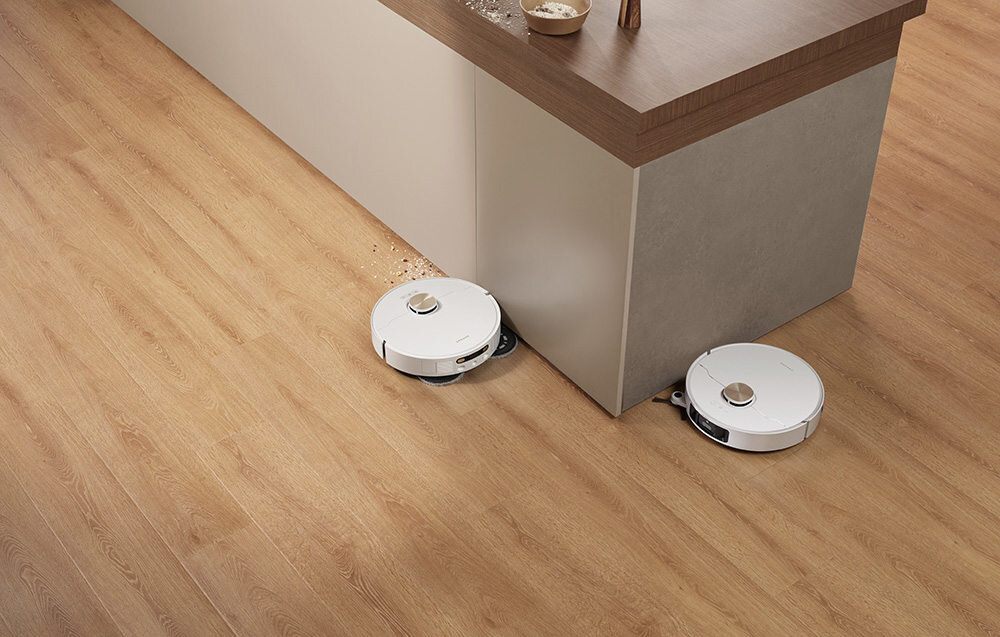 Dwa białe roboty sprzątające poruszają się po drewnianej podłodze obok kuchennej wyspy, zbierając rozsypane okruchy. Roboty mają okrągły kształt i są w trakcie pracy.