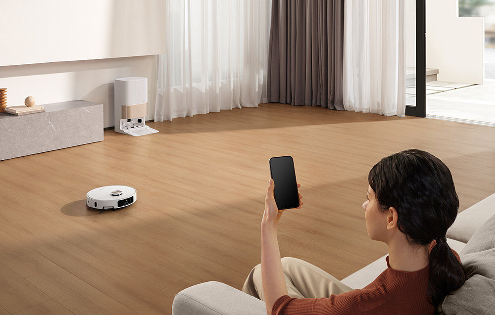 Osoba siedzi na kanapie, trzymając smartfon, podczas gdy robot sprzątający porusza się po drewnianej podłodze. W tle widać stację dokującą przy ścianie oraz zasłony przy dużym oknie.