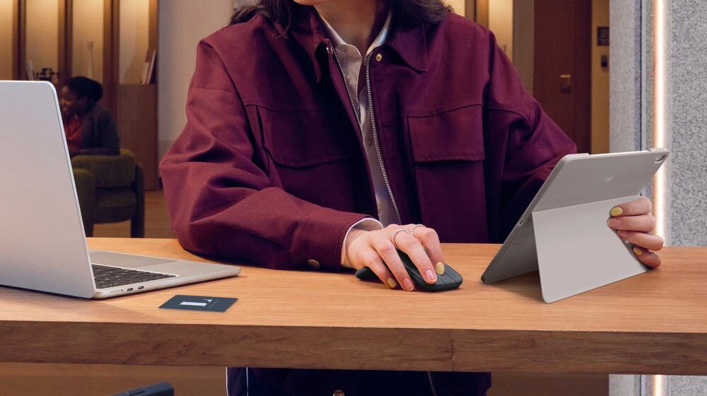LOGITECH Mobi Fold Składana konstrukcja Rozłożenie urządzenia automatycznie je włącza złożenie wyłączenie - Dłoń obsługująca płaską mysz na blacie roboczym podczas pracy z tabletem opartym o podstawkę i komputerem przenośnym.