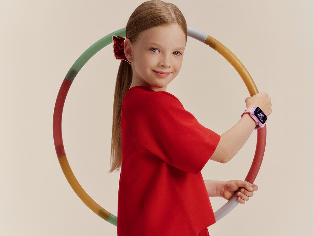 Dziewczynka w czerwonym ubraniu trzyma kolorowe koło i ma na nadgarstku smartwatch. Smartwatch jest w kolorze różowym z wyświetlaczem pokazującym kolorowy obraz.