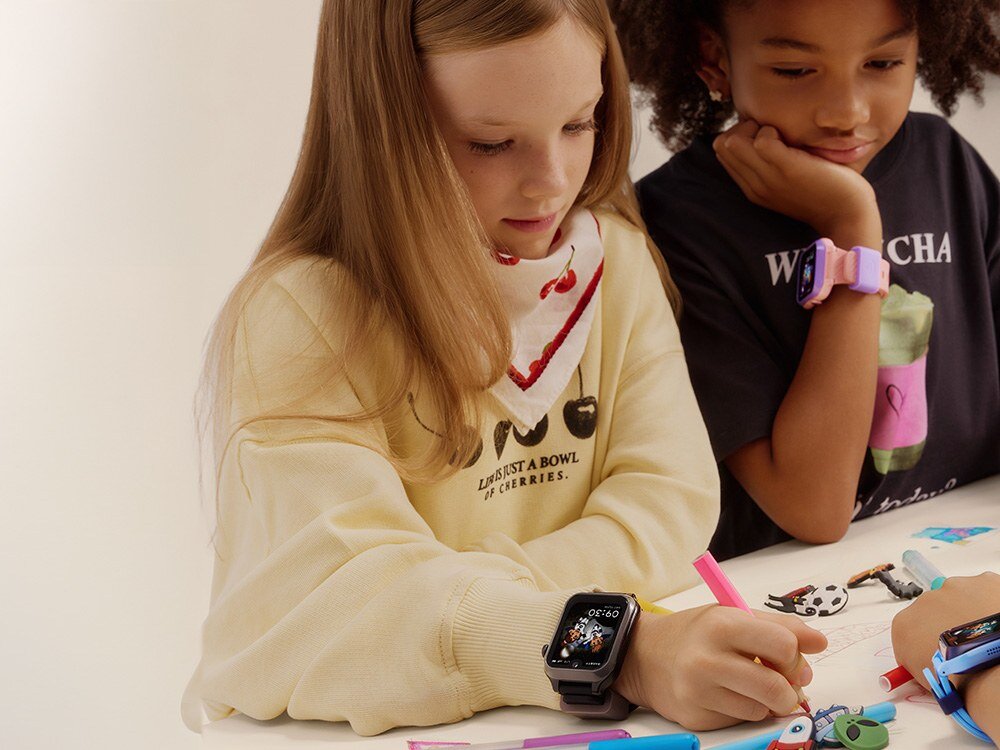 Dwoje dzieci siedzi przy stole, jedno z nich ma na nadgarstku smartwatch z kolorowym wyświetlaczem. Na stole leżą kolorowe pisaki i drobne zabawki.