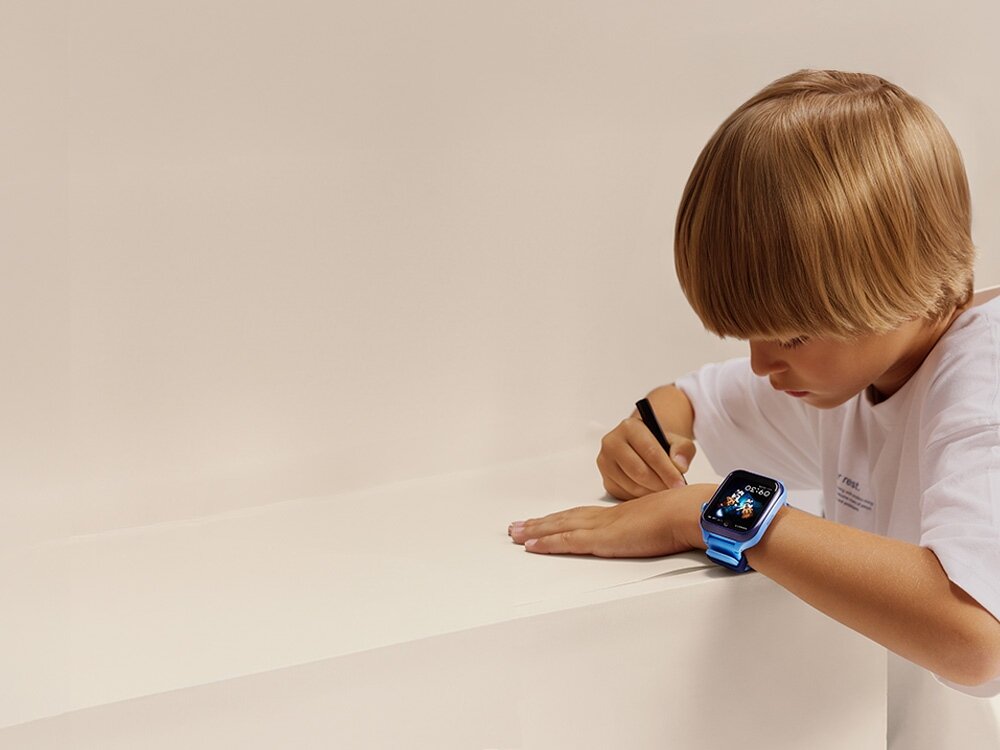 Dziecko w białej koszulce pisze na jasnej powierzchni, mając na nadgarstku niebieski smartwatch z kolorowym wyświetlaczem.