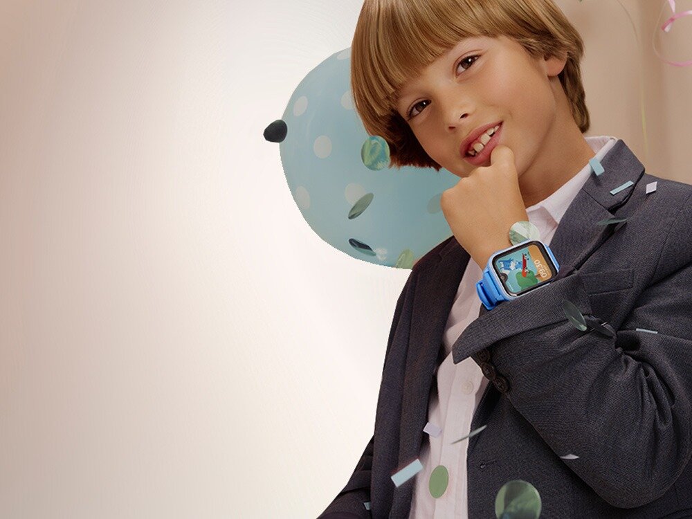 Dziecko w garniturze prezentuje smartwatch na nadgarstku, z kolorowym wyświetlaczem. W tle widoczny jest balon w kropki i konfetti.