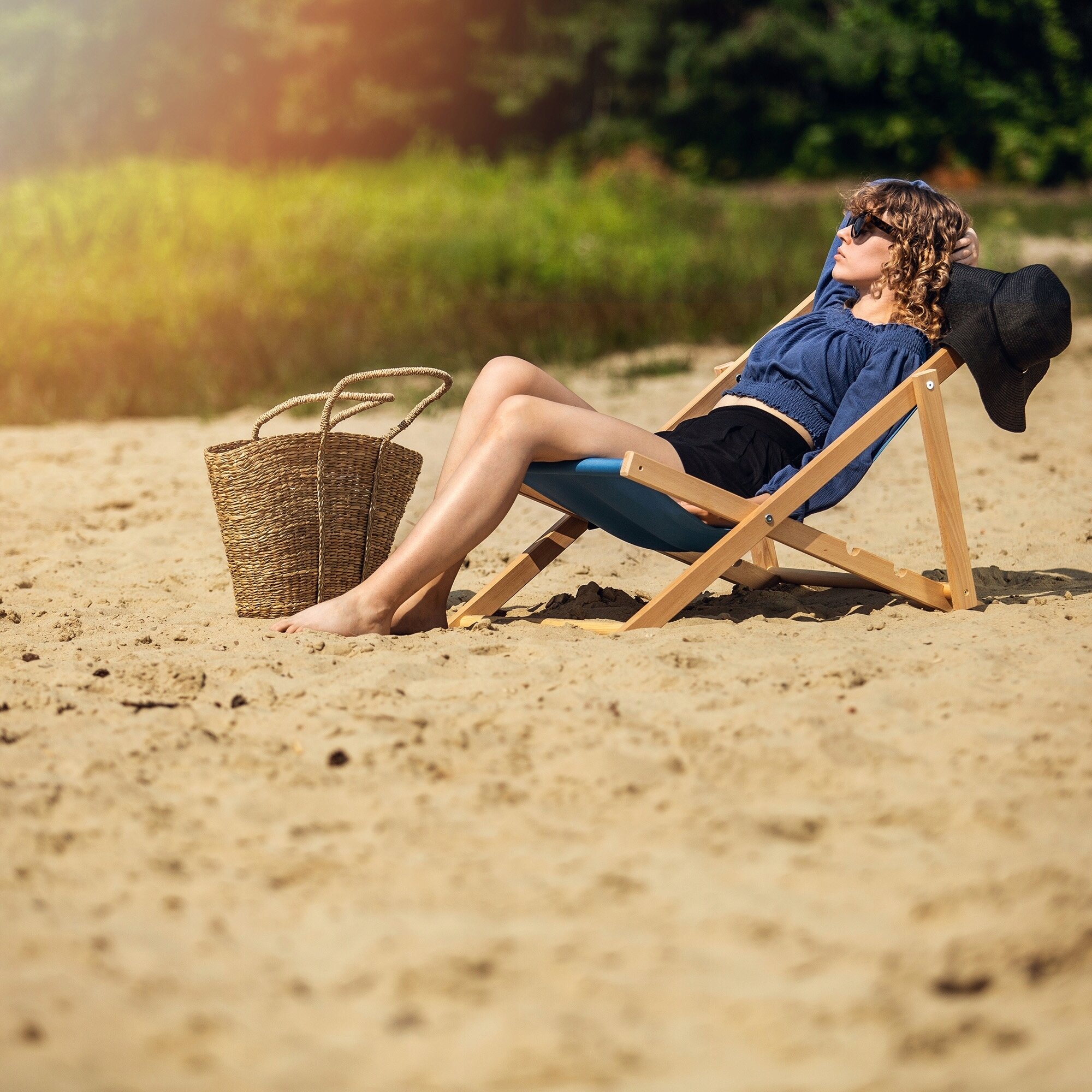 Osoba siedzi na drewnianym, składanym leżaku z niebieskim materiałem na piaszczystej plaży. Obok leżaka znajduje się pleciona torba.