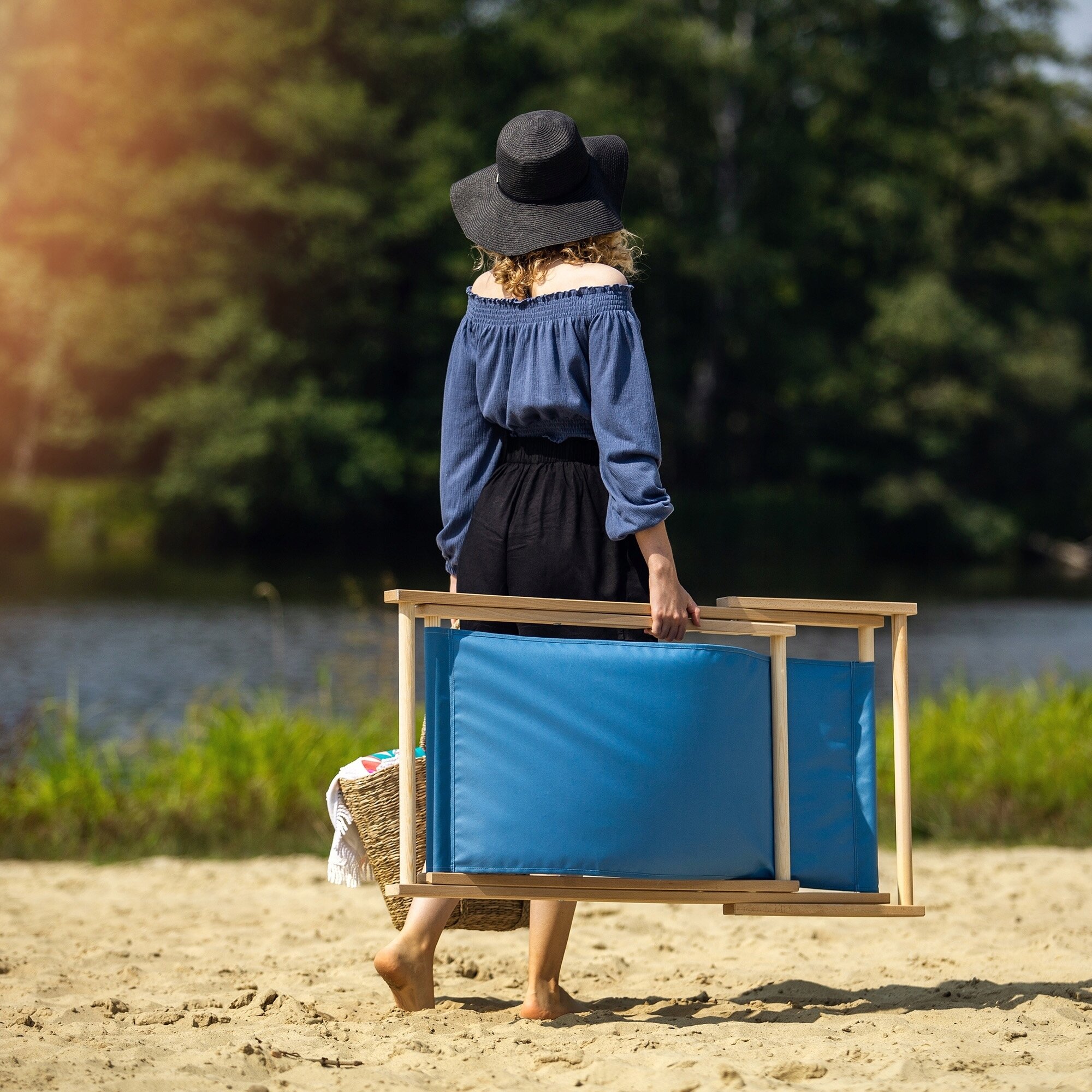 Osoba na plaży niesie składany leżak z niebieskim materiałem i drewnianą ramą. W tle widać wodę i drzewa.