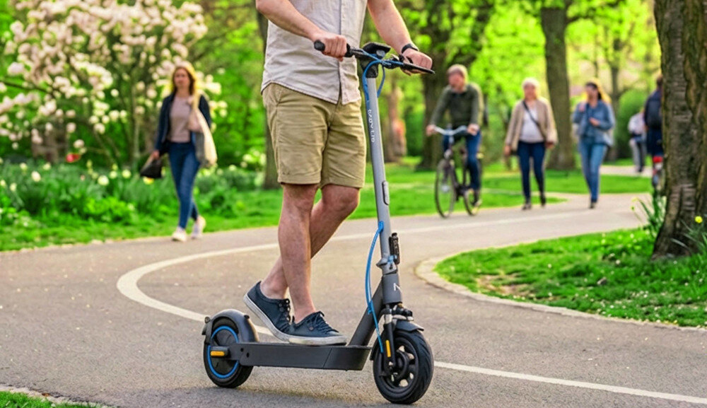 Osoba jedzie na szaro-turkusowej hulajnodze elektrycznej po asfaltowej ścieżce w parku. W tle widać zieleń drzew i kilka osób spacerujących lub jadących na rowerach.