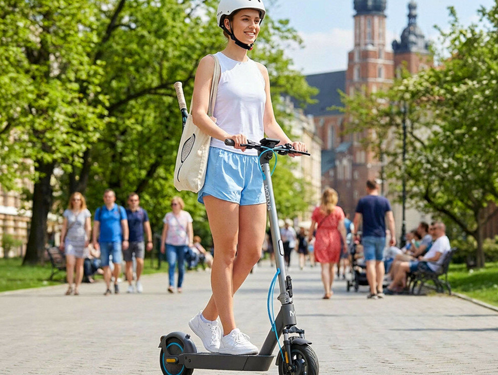 Osoba w kasku jedzie na szaro-turkusowej hulajnodze elektrycznej po parku. W tle widać spacerujących ludzi i drzewa.