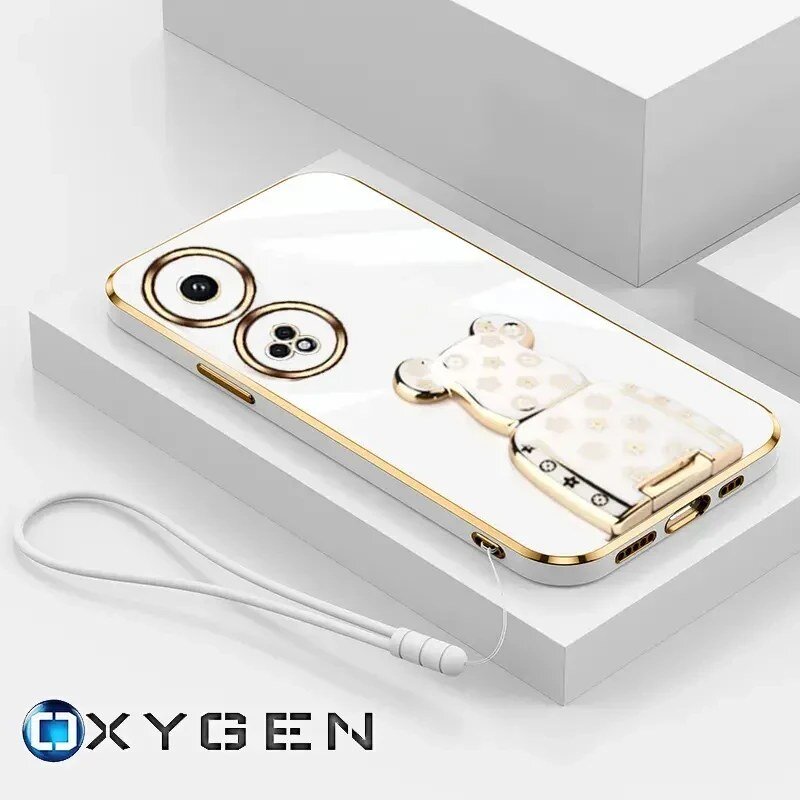 Na obrazie widoczne jest białe etui na telefon z ozdobnym uchwytem w kształcie misia i złotymi elementami. W dolnej części znajduje się logo \'OXYGEN\'.