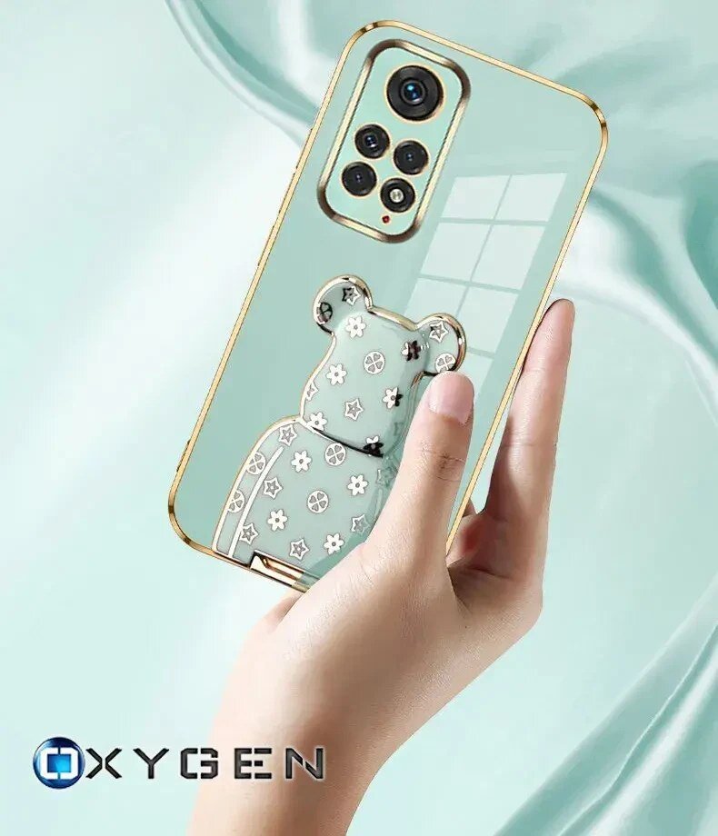 Osoba trzyma przezroczyste etui na telefon z ozdobą w kształcie misia. Na dole widoczne jest logo \'OXYGEN\'.