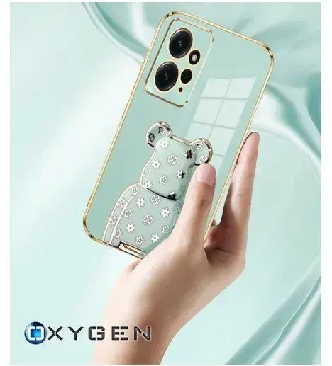 Osoba trzyma telefon w przezroczystym etui z ozdobnym motywem misia. Na dole widoczne jest logo OXYGEN.