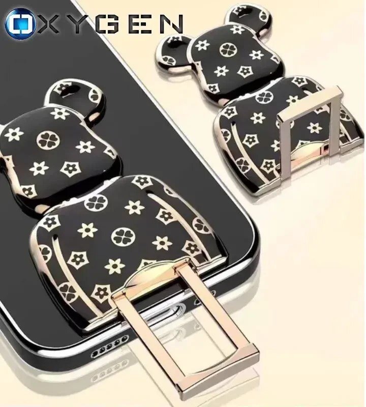Na obrazie widoczne jest czarne etui na telefon z ozdobnym uchwytem w kształcie misia, pokrytym wzorem kwiatowym. W lewym górnym rogu znajduje się logo \'OXYGEN\'.
