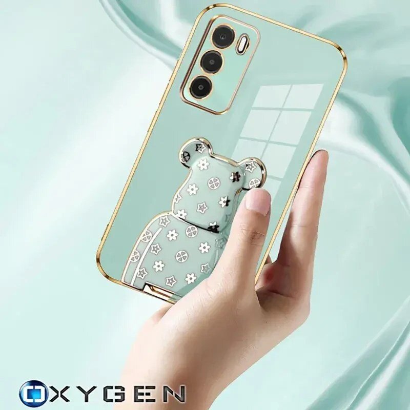 Osoba trzyma przezroczyste etui na telefon z ozdobnym uchwytem w kształcie misia, pokrytym wzorami kwiatów i symboli pokoju. W dolnym rogu widoczne jest logo OXYGEN.