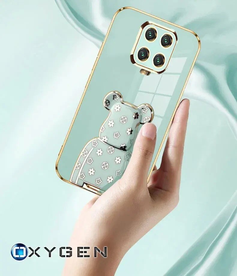 Osoba trzyma przezroczyste etui na telefon z ozdobnym motywem misia i złotymi krawędziami. W lewym dolnym rogu widoczne jest logo \'OXYGEN\'.