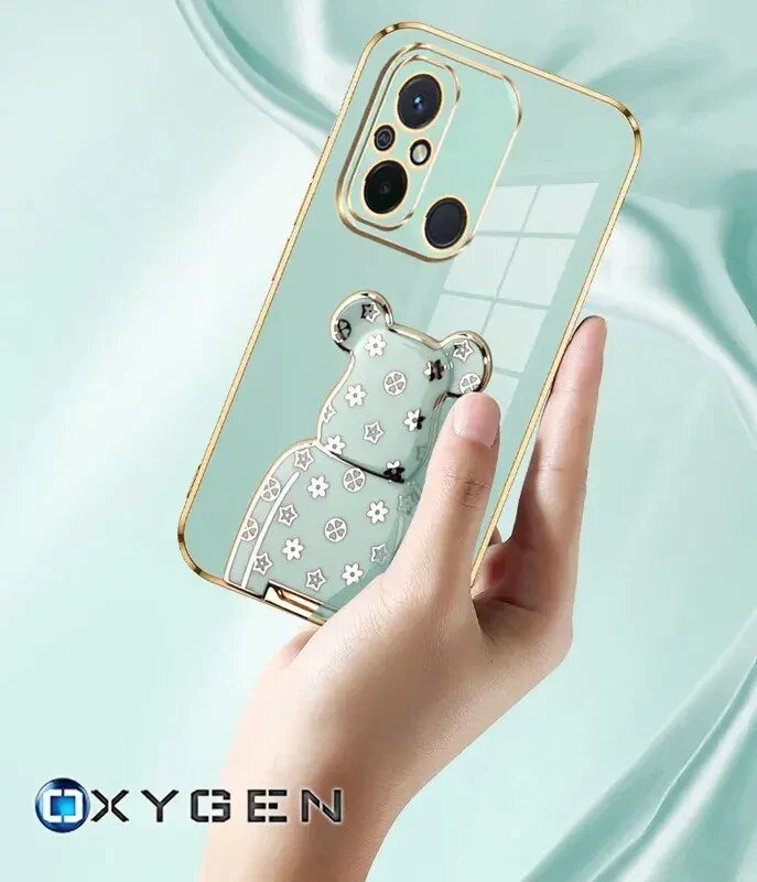 Osoba trzyma przezroczyste etui na telefon z ozdobnym motywem misia i złotymi akcentami wokół aparatu. W lewym dolnym rogu widoczne jest logo \'OXYGEN\'.