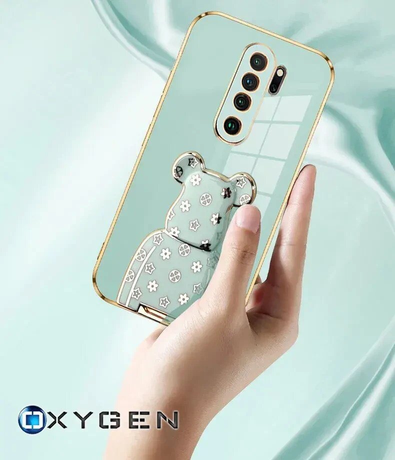Osoba trzyma telefon w etui z motywem misia ozdobionego kwiatkami i gwiazdkami. Na dole widoczne jest logo \'OXYGEN\'.