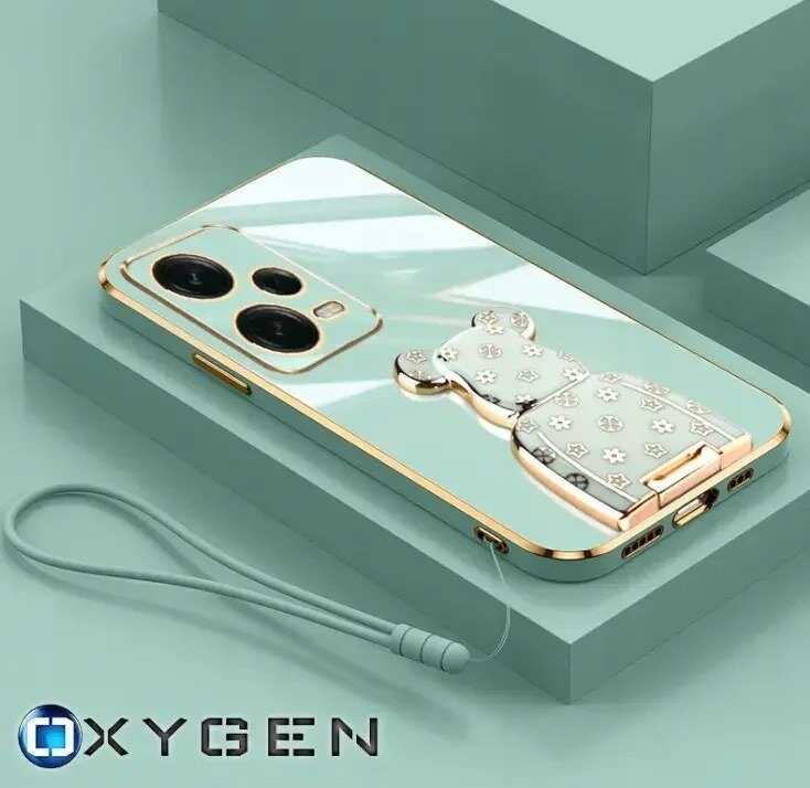 Na obrazie widoczne jest etui na telefon z metalicznym wykończeniem i ozdobnym uchwytem w kształcie misia. W dolnym rogu znajduje się logo \'OXYGEN\'.