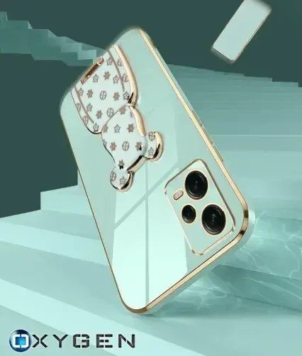 Etui na telefon z metalicznymi krawędziami i ozdobnym uchwytem w kształcie misia, na tle schodów. Widoczne logo: OXYGEN.